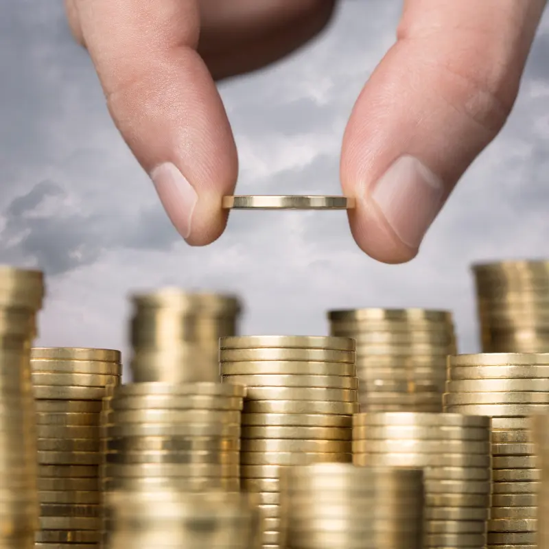 Symbolbild: Mehrere Stapel goldener Geldmünzen. Man sieht, wie eine Hand eine Münze obendrauf legt.