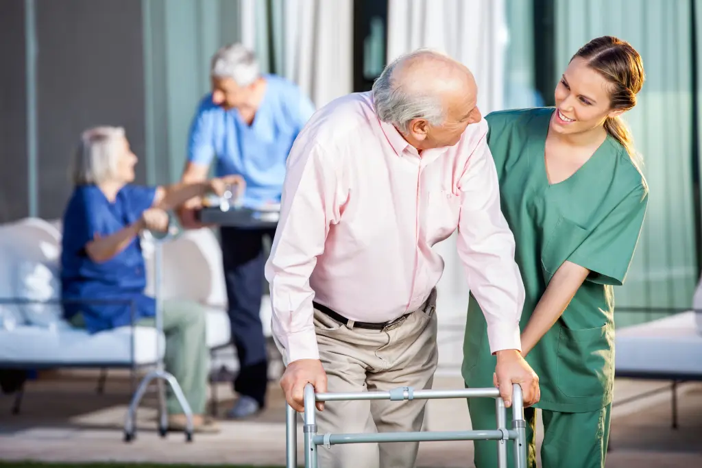 Foto: Ein älterer Mann am Rollator wird von einer jungen Pflegerin durch eine Pflegeeinrichtung geführt, im Hintergrund sitzen ebenfalls zwei ältere Menschen.