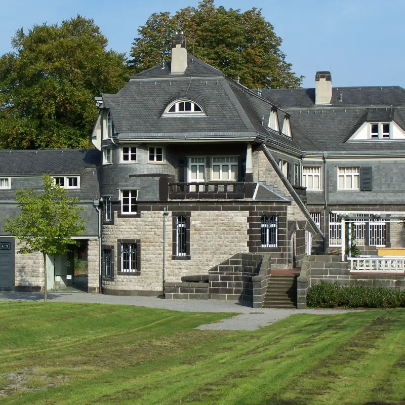 Foto: Eine große schöne Villa mit dunklem Dach, im Vordergrund ein gut gepflegter Rasen.