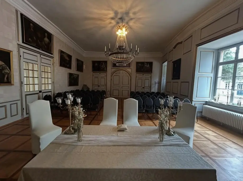 Foto: Ein breiter Tisch mit samtener Tischdecke und mehreren Stühlen in einem Festsaal.