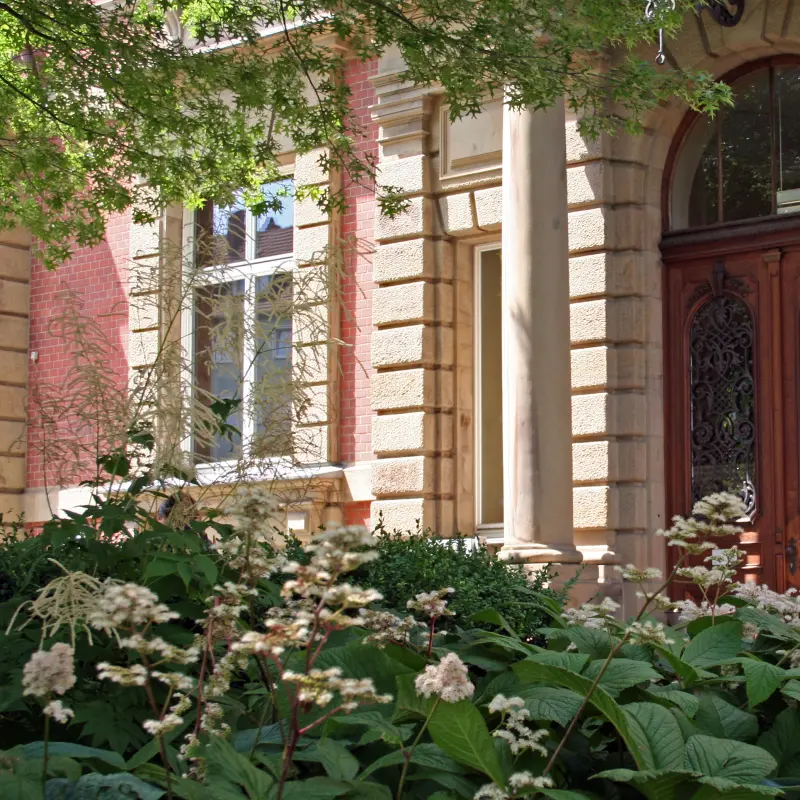 Foto: Der Eingang einer Villa mit einer hölzernen Tür mit aufwendigen Verzierungen zwischen zwei sandfarbenen Säulen.