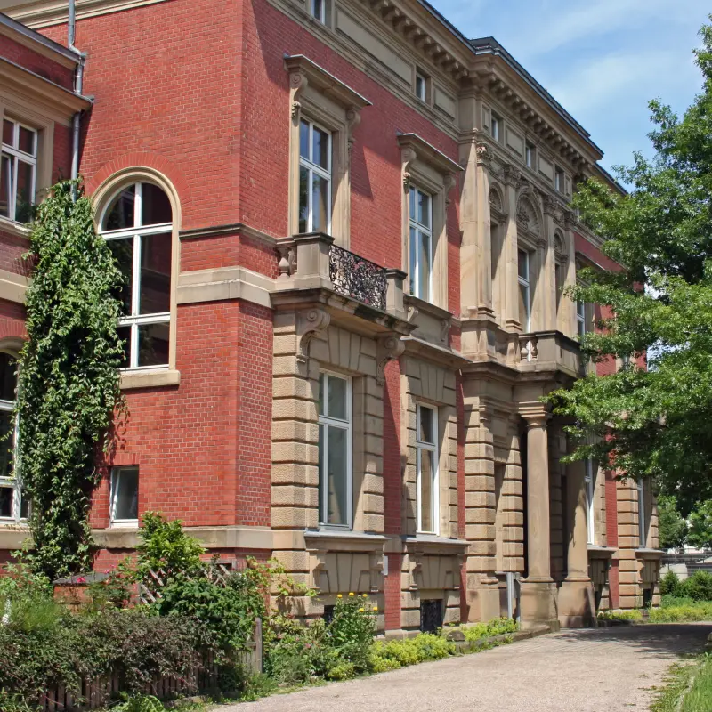 Foto: Die schräge Seitenansicht einer Villa mit roter Fassade mit beigen Stein-Elementen, rechts im Bild befindet sich ein Baum.