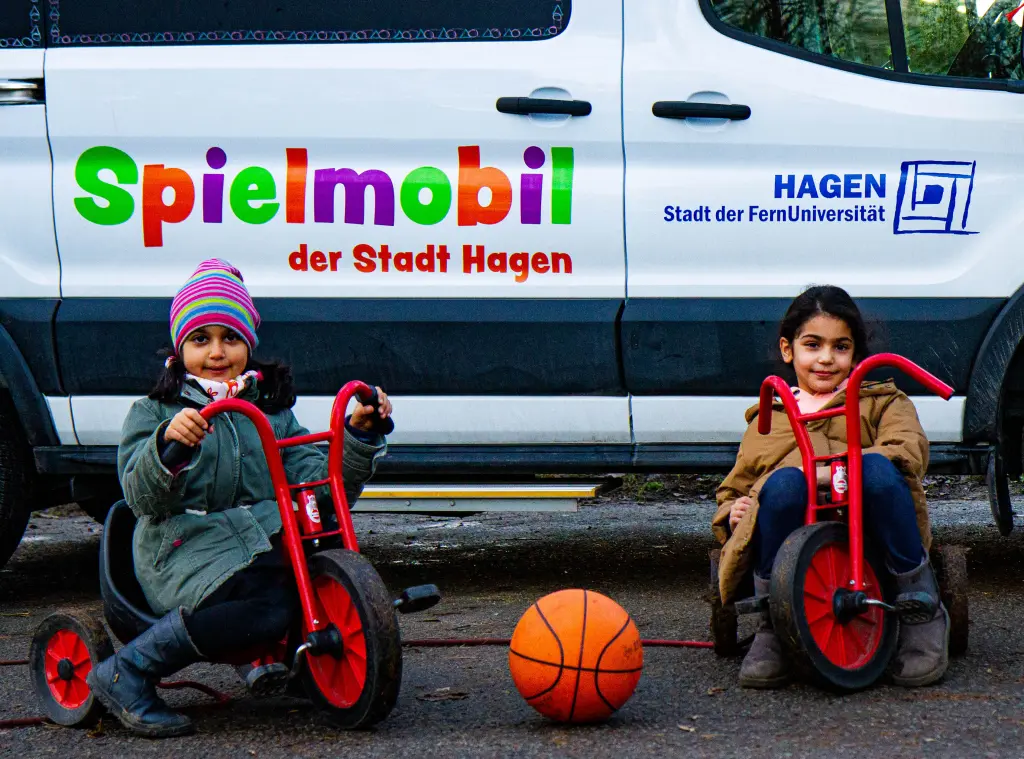 Foto: Das Fahrzeug des Spielmobils geparkt auf einer Straße, davor sitzen zwei Kinder in Dreirädern.