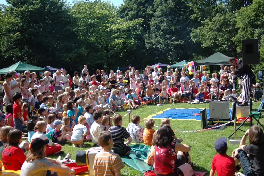 Foto: Eine große Gruppe von Kindern sitzt auf einer Wiese und guckt einer Zirkus-Veranstaltung zu.