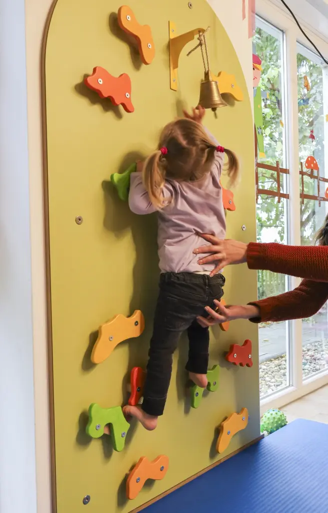 Foto: Ein Kleinkind klettert mit Unterstützung eine altersgerechte Spielwand hoch.