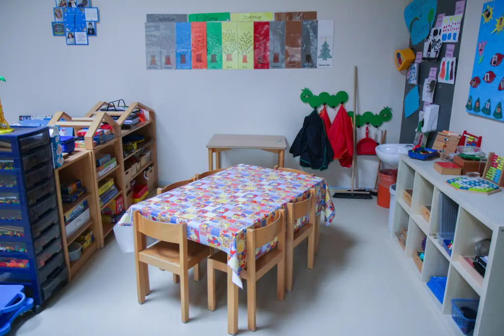 Foto: Ein Raum in einer Kindertagesstätte mit einem Bücherregal, vielen Postern an den Wänden und einem gedeckten Tisch.