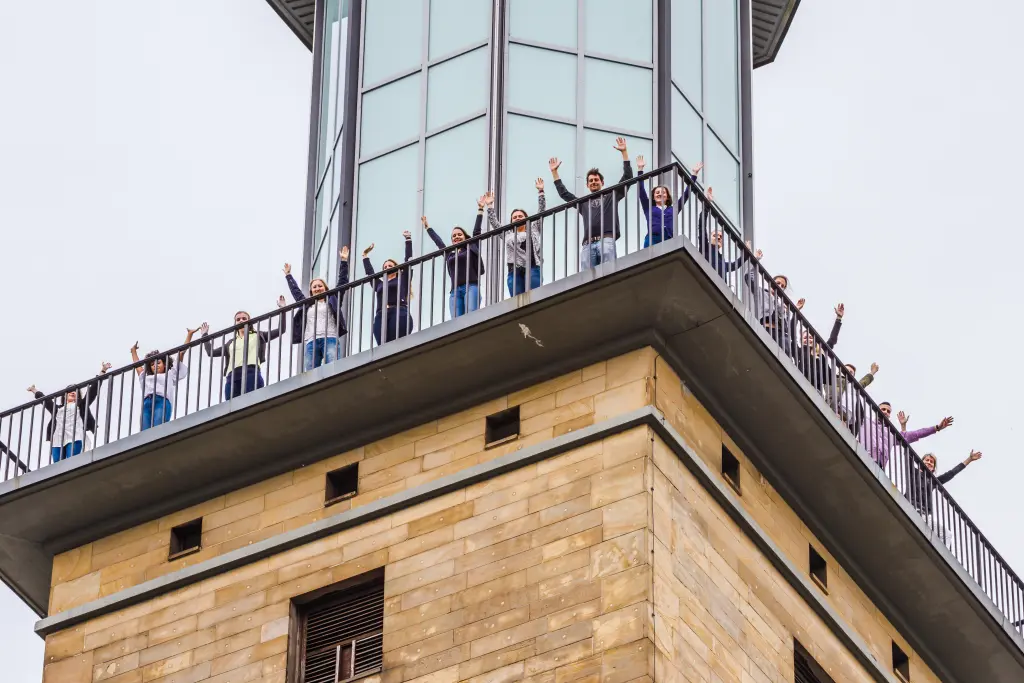 Foto: Eine Gruppe Auszubildener steht auf dem Rathausturm der Stadt Haben und hebt die Hände.